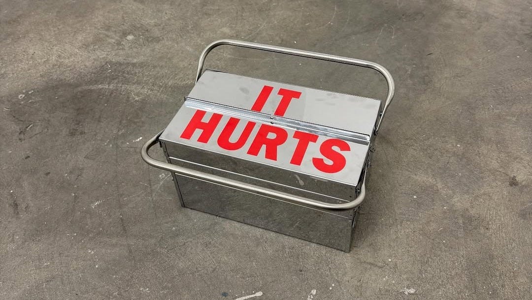 A photo of a silver metal box on a concrete floor. In bold red letters, text says, It hurts.