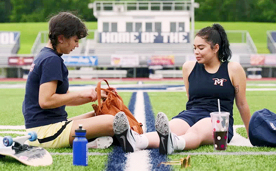 gif do filme 'crush'. as protagonistas estão sentadas num campo de esporte da escola