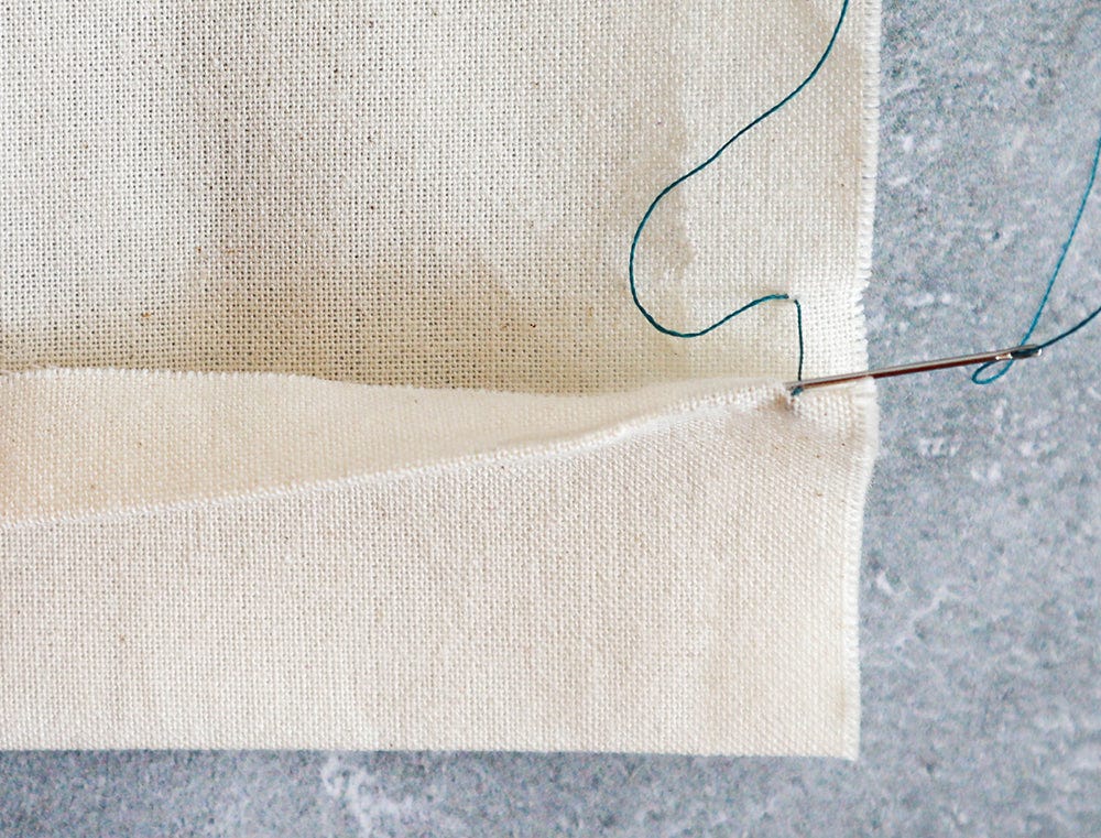 Cream muslin fabric with hem folded up, needle with teal thread sewing a slip stitch