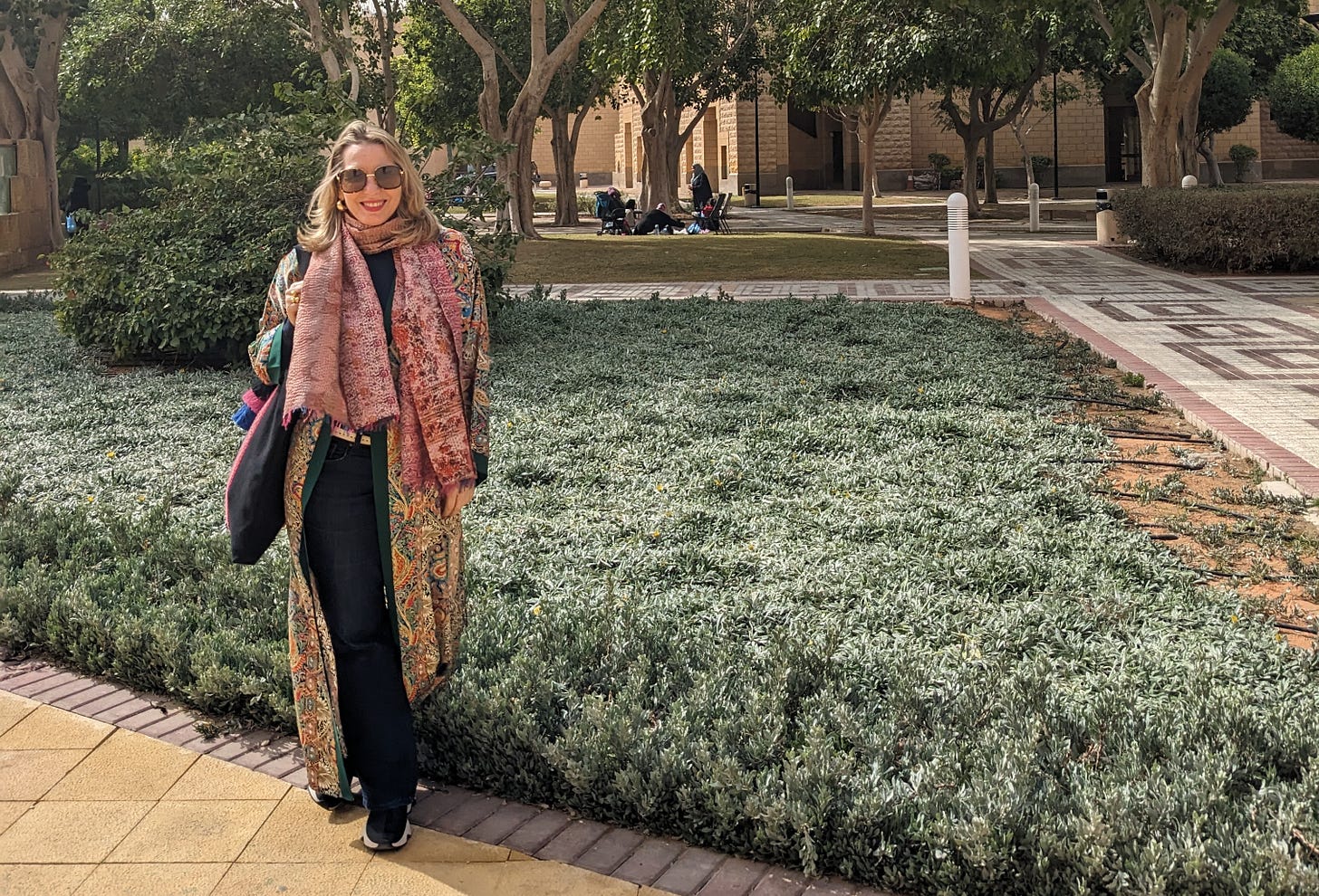 White woman wearing a multi-colored robe and scarf, standing in a garden. She is smiling and wearing sunglasses. 