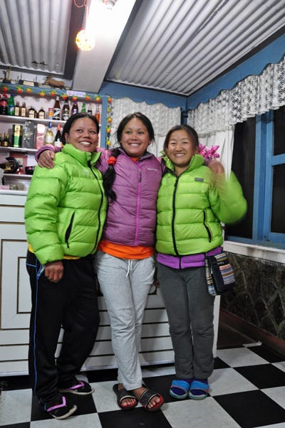 Three Nepali women wearing green and pink down coats, hugging and smiling at the camera. 