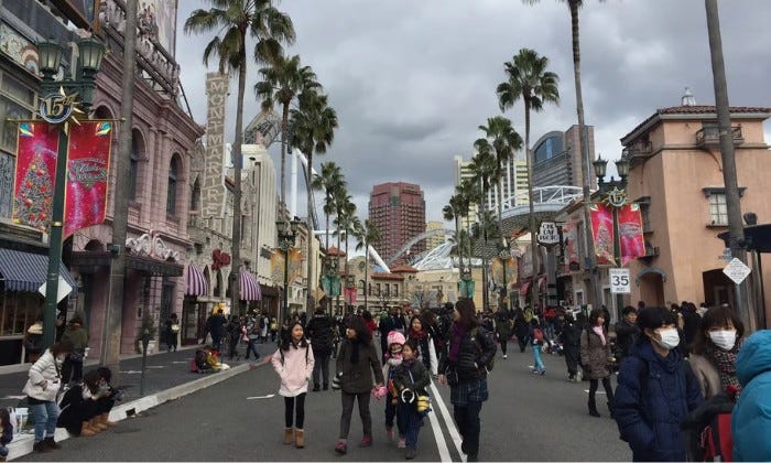 Palm-lined street at Universal Studios Japan, flanked by ornate Hollywood-style facades and holiday banners. Crowds in winter coats walk the road under a cloudy sky, with the city skyline visible in the background.