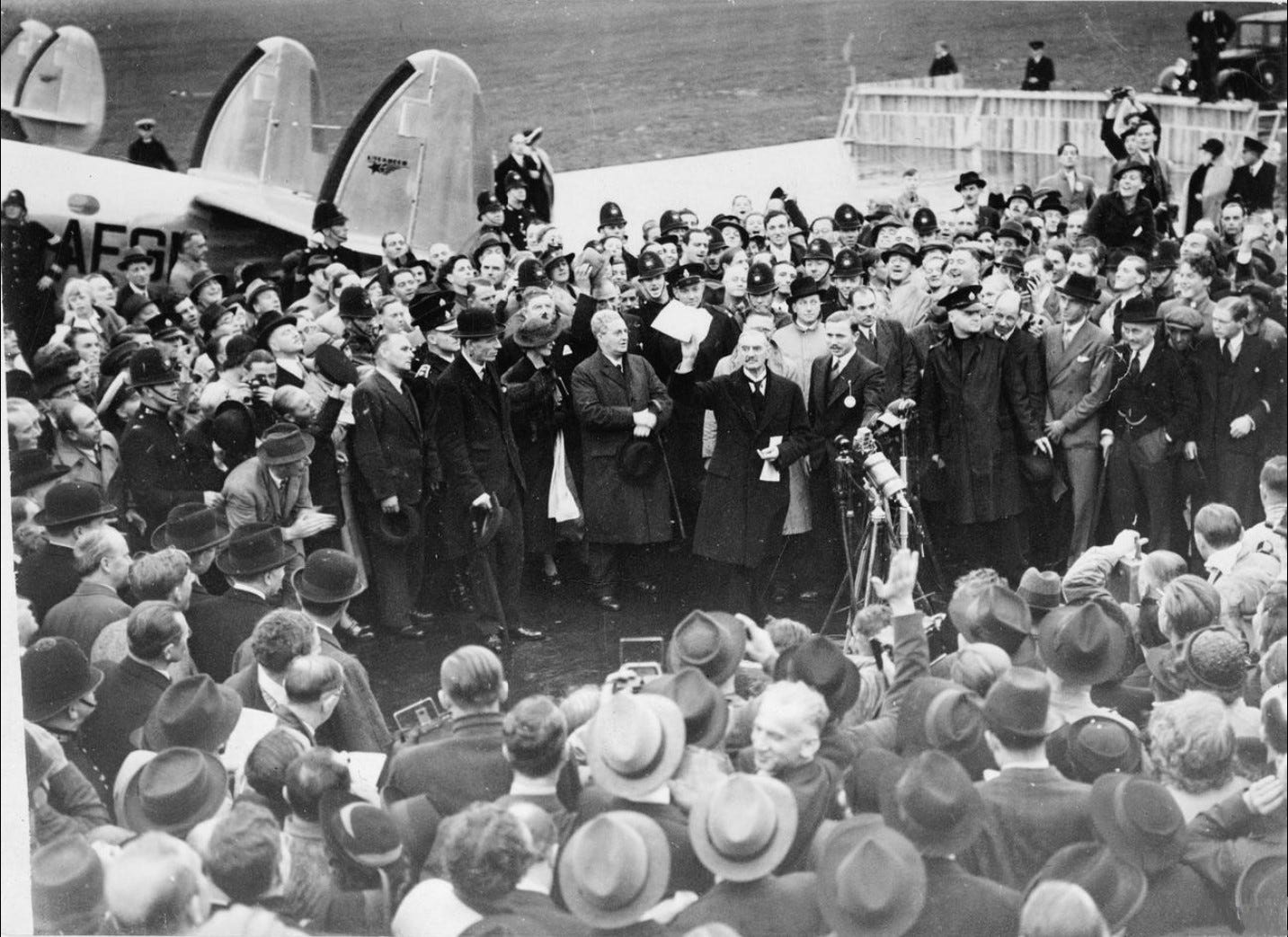 A large crowd on an airfield; British prime minister Neville Chamberlain presents an assurance from German chancellor Adolf Hitler. A large crowd on an airfield; British prime minister Neville Chamberlain presents an assurance from German chancellor Adolf Hitler.