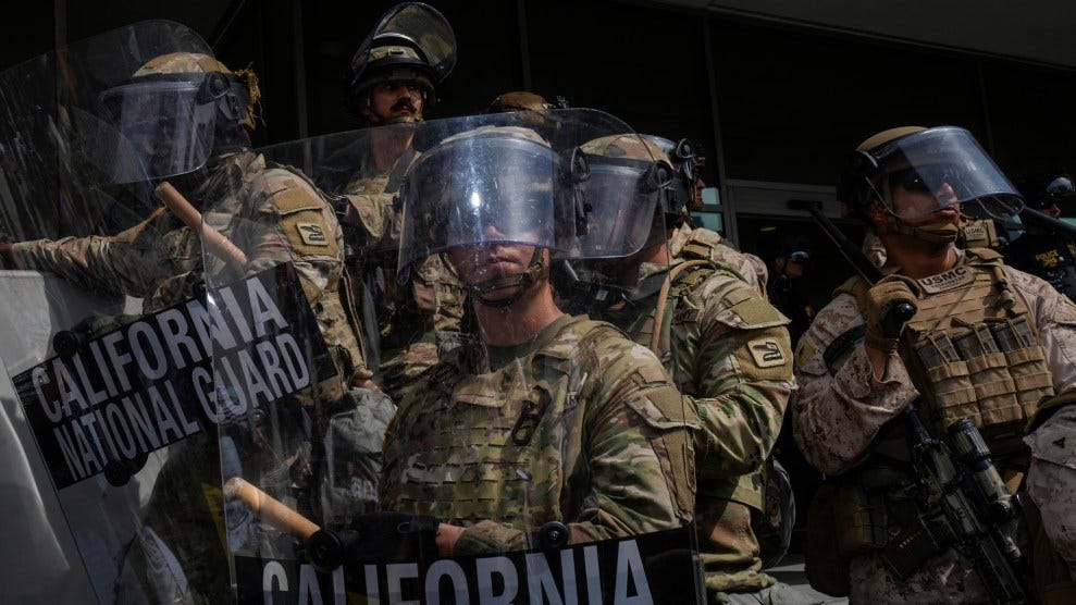 Several rows of uniformed troops stand on alert. They are dressed in camouflage fatigues and wear helmets as they hold protective clear shields that read “California National Guard.”