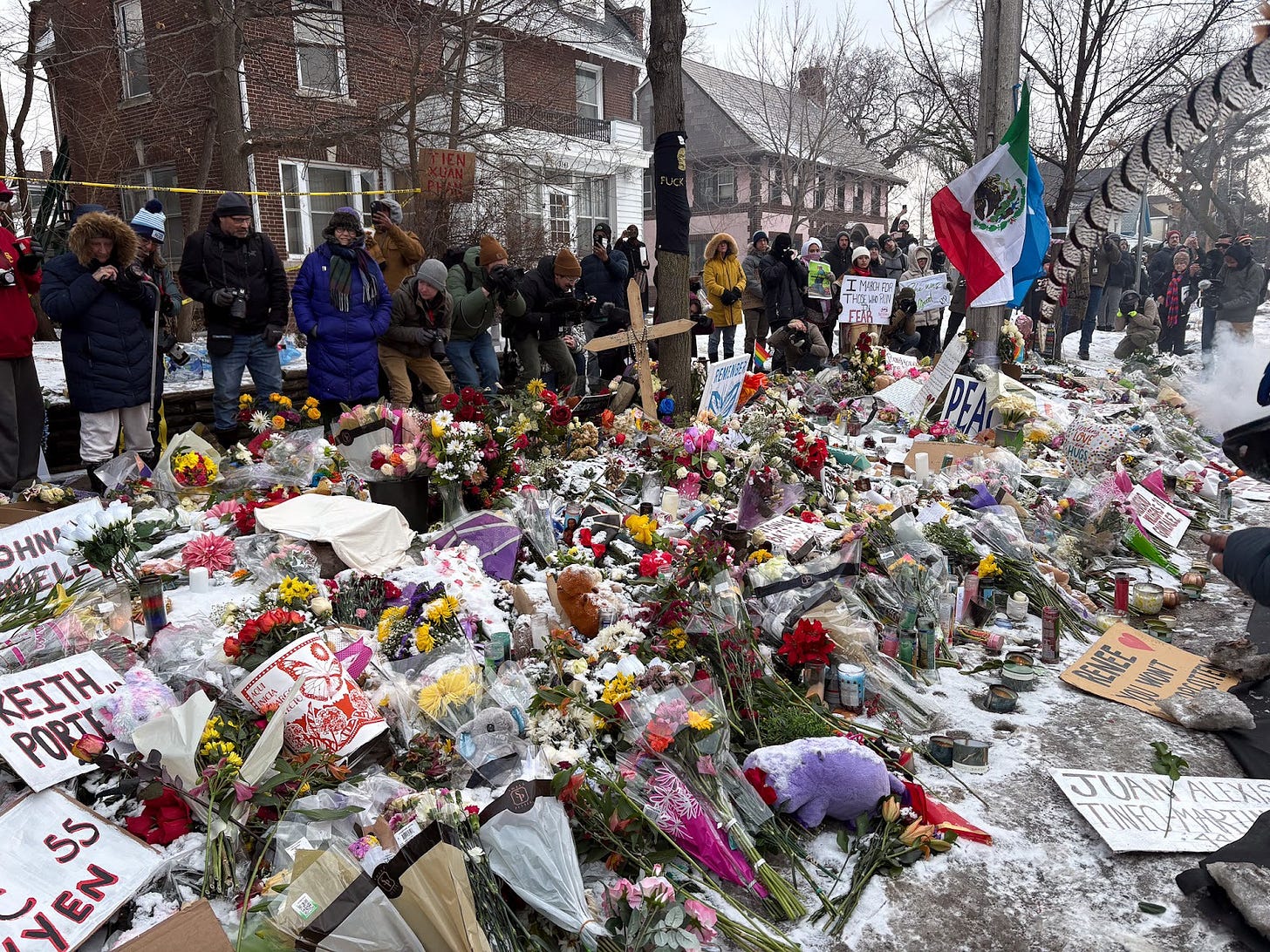 A crowd stands behind a pile of flowers and signs memorializing victims of ICE brutality