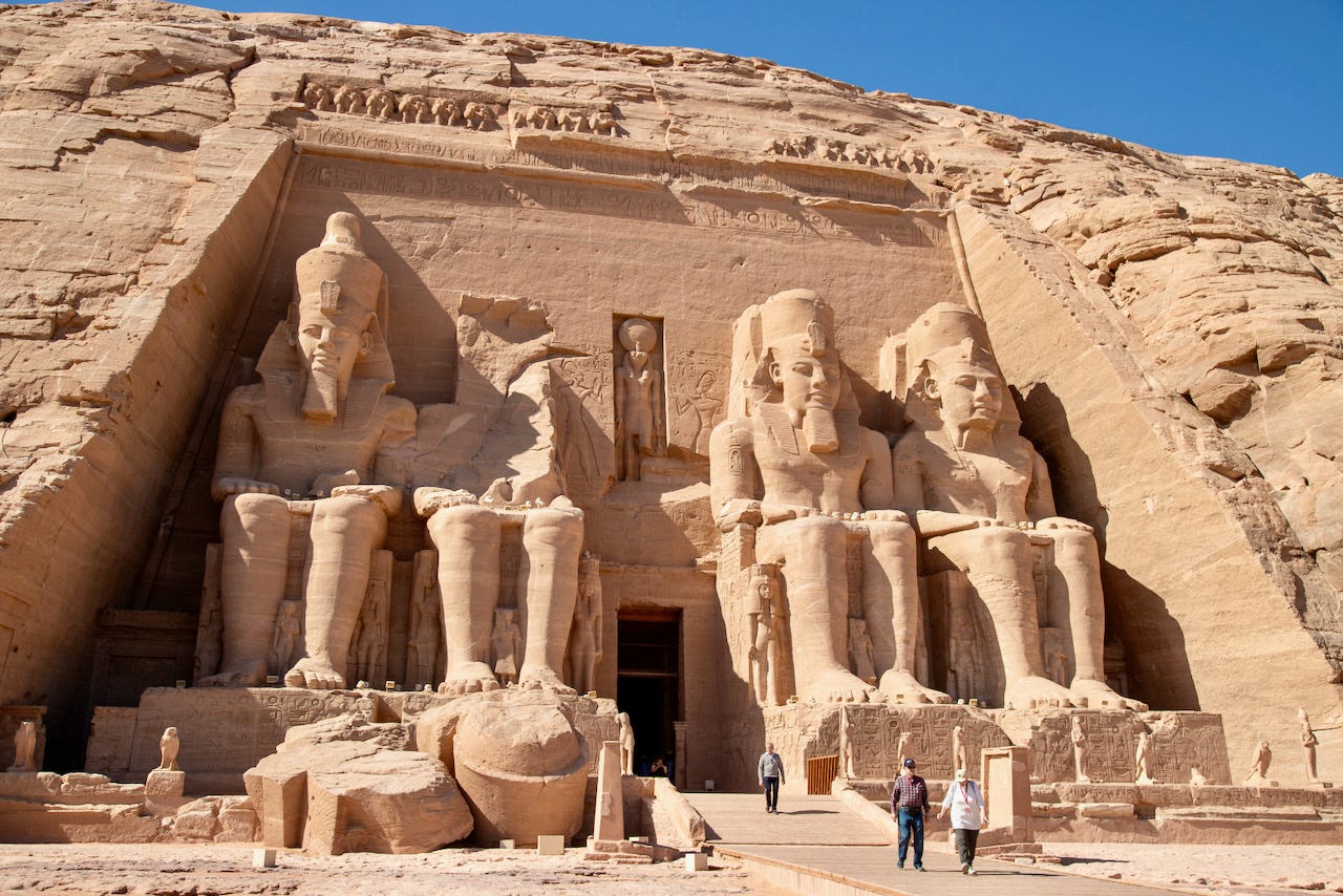 The facade of the Great Temple at Abu Simbel with four colossal seated statues of Ramesses II carved into the sandstone cliff face, with visitors walking at the base for scale
