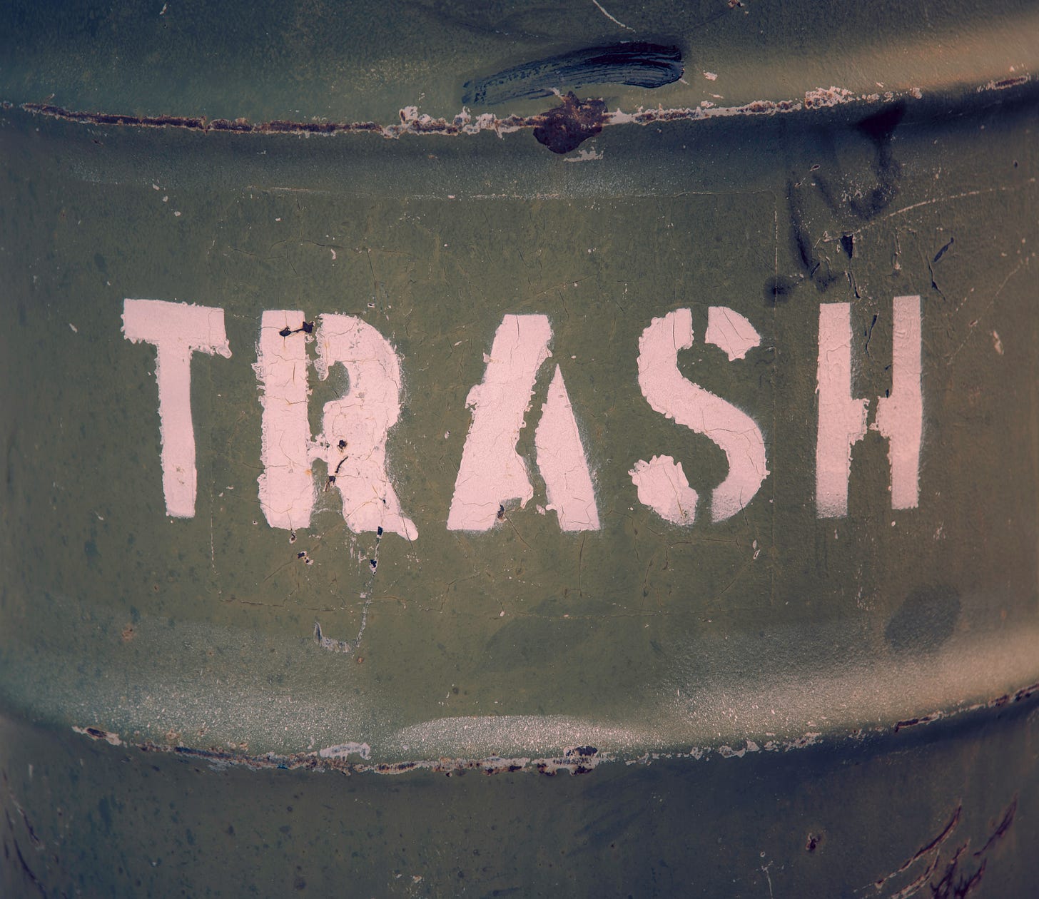 Weathered green metal trash container with faded pink stenciled 'TRASH' lettering, showing signs of rust and wear
