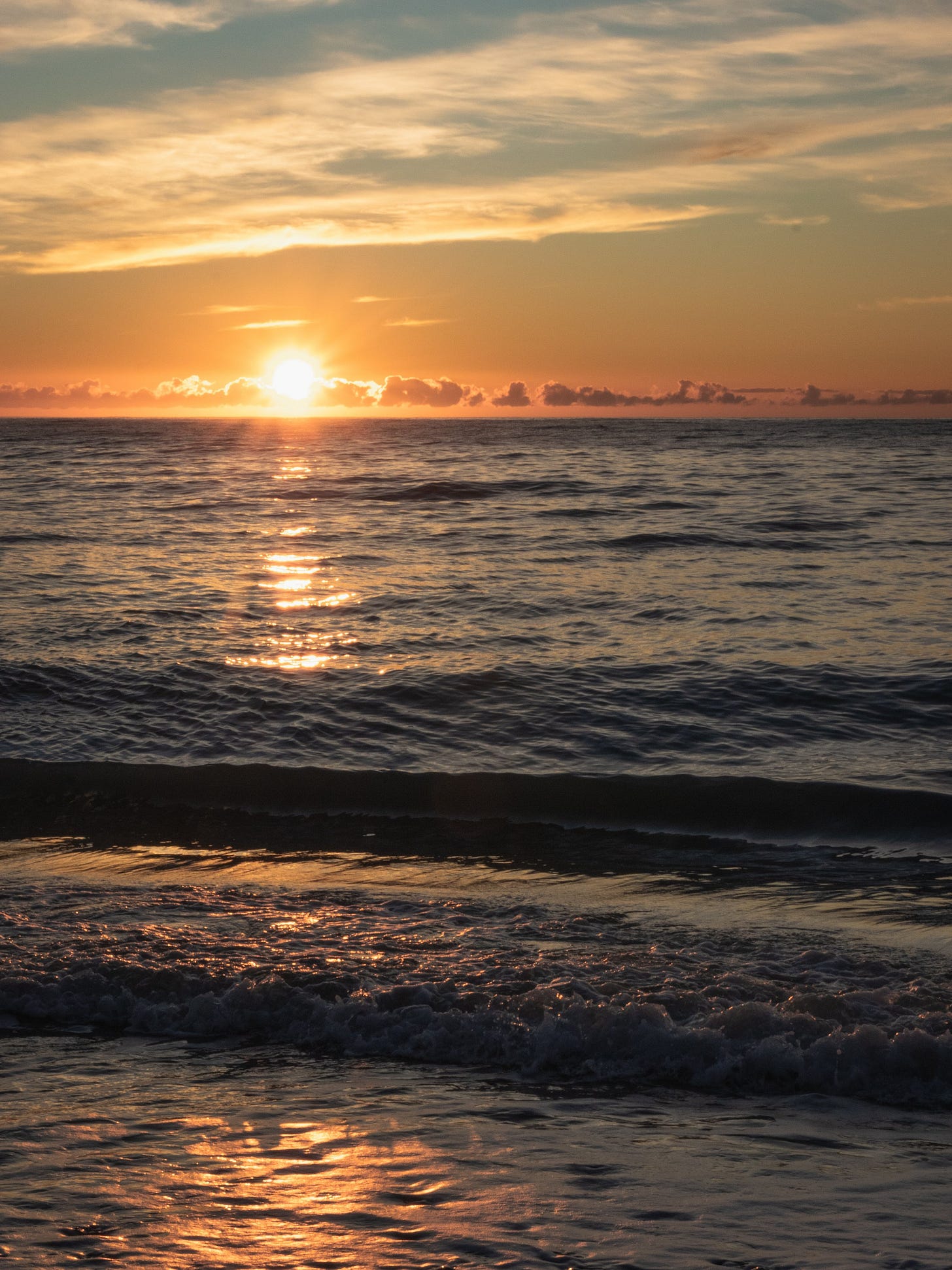 A portrait scene of sunrise at the beach. The ocean is smooth and a small wave is about to land on the beach.