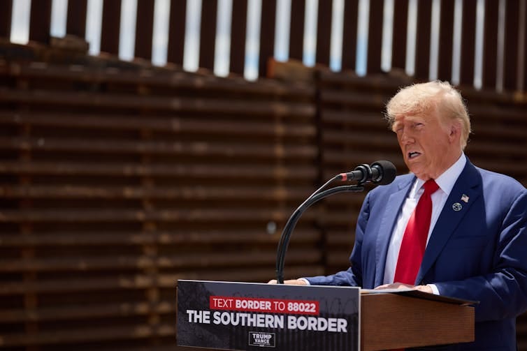 Trump speaking at a podium that reads 'the southern border', with a large border fence in the background