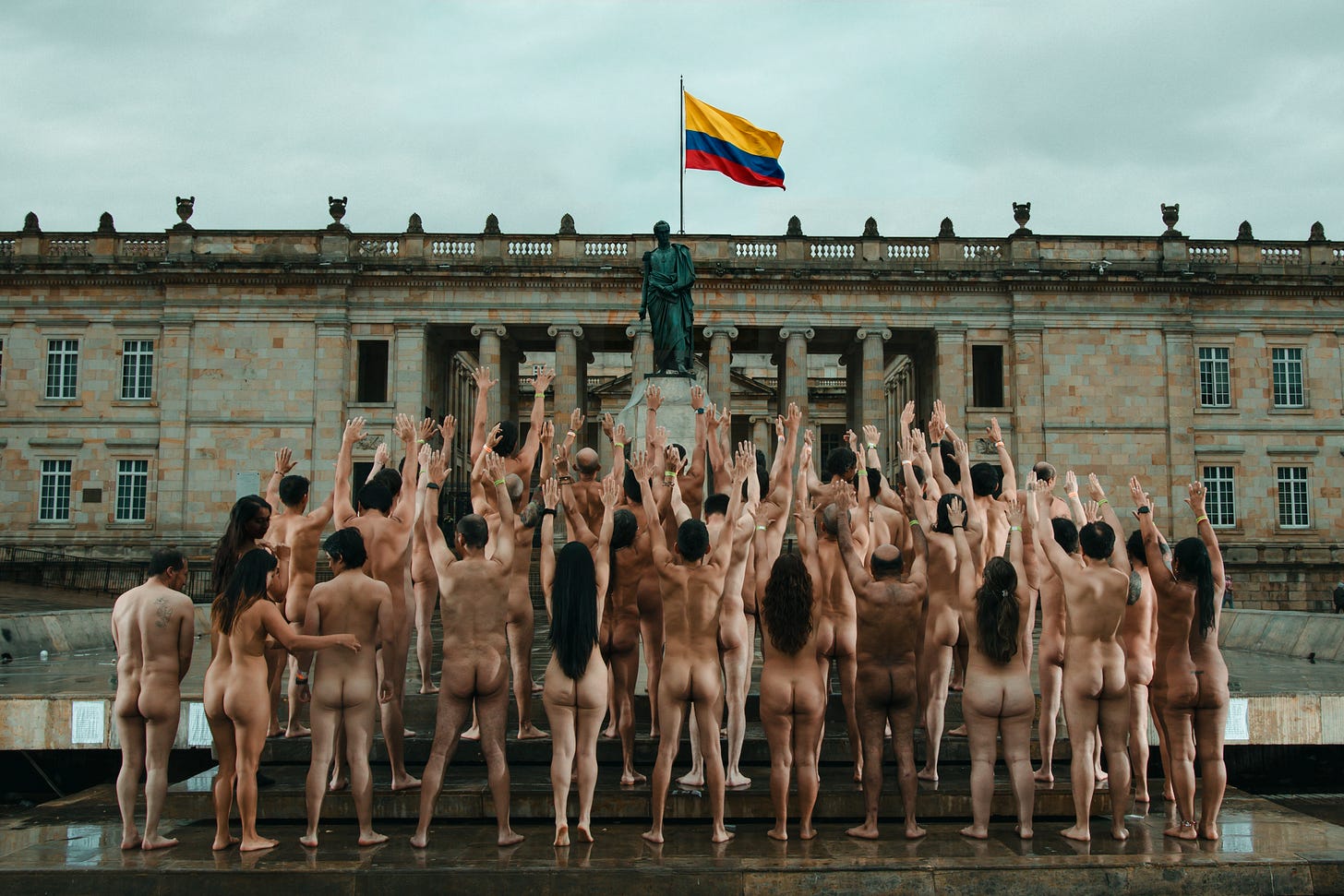 A large group of nude people stands with arms raised on the wet stone of Plaza de Bolívar, facing the statue of Simón Bolívar and the columned facade of Colombia's Capitolio Nacional, with the Colombian flag flying overhead under an overcast sky. A large group of nude people stands with arms raised on the wet stone of Plaza de Bolívar, facing the statue of Simón Bolívar and the columned facade of Colombia's Capitolio Nacional, with the Colombian flag flying overhead under an overcast sky.