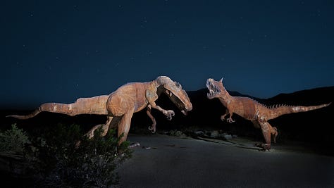 five photos that Kirk Marshall took. 1. B&W rolling sand dunes. 2. two metal dinosaur sculptures in a mock fight against a dark desert night. 3. B&W cactus along a road with the shadow of one across the road. 4. B&W an old destroyed tyre showing the decayed side walls while still attached to a rusted bumper. 5. A metal dragon sculpture created to look like it’s underneath the surface of the earth. A bright blue sky and some low lying hazy hills in the background.
