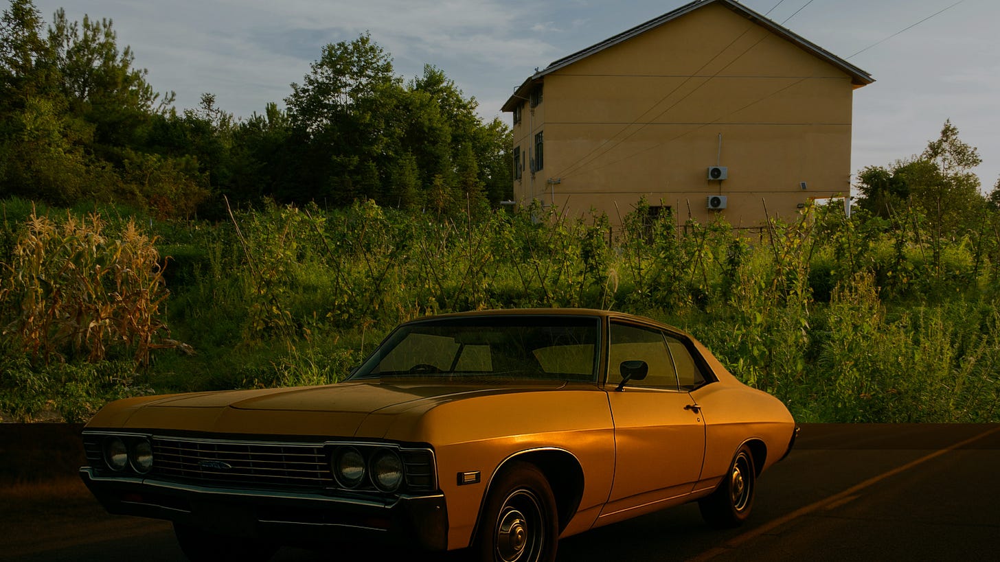 A vintage yellow Impala parked near a cornfield and house at dusk, its windows darkened and chrome catching the morning light