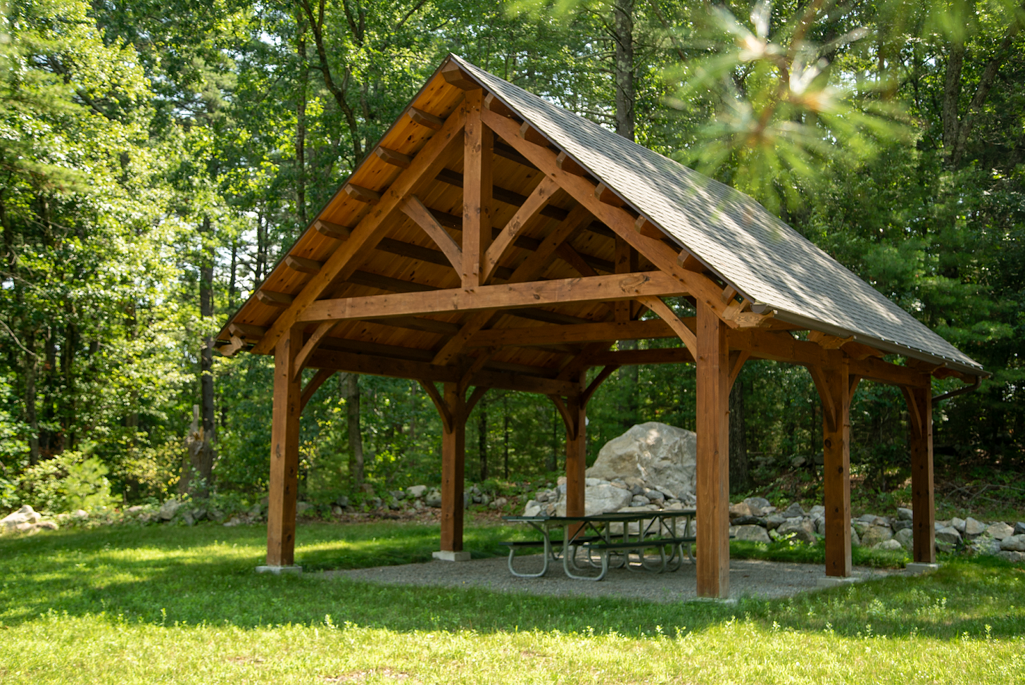 A wooden roofed pavilion in front of a wooded area on a grassy lawn. Picnic tables are under the cabin.