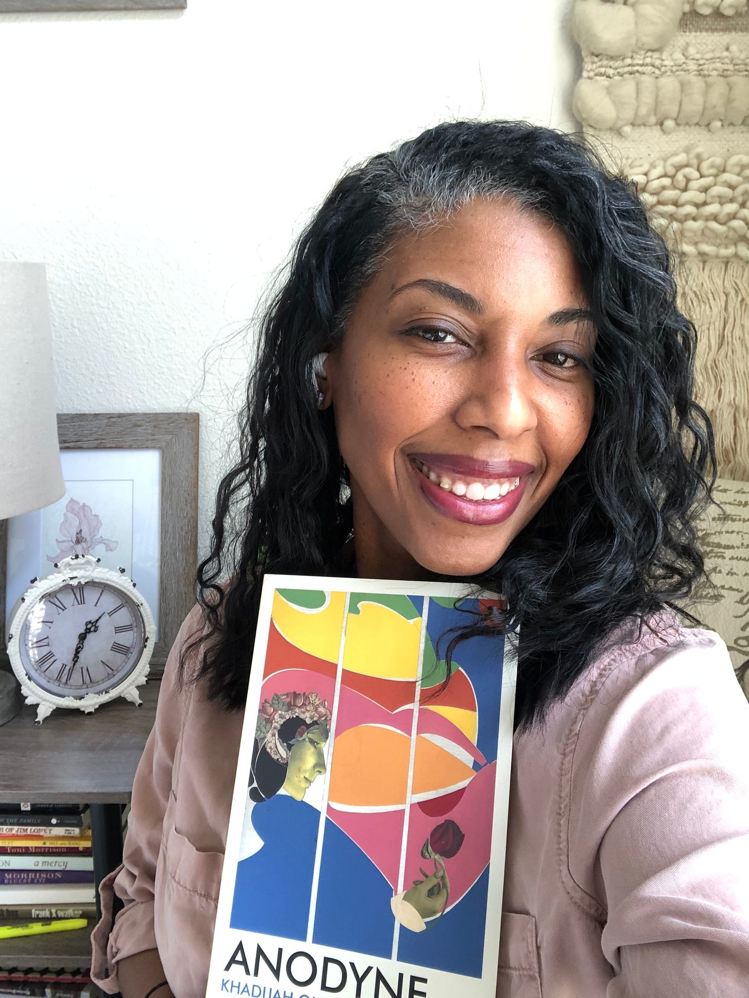 Khadijah with black wavy hair, gray showing at the roots by about an inch and a half. She is holding a book and smiling. The backdrop is a corner with a clock, a lamp, and a beige tapestry.