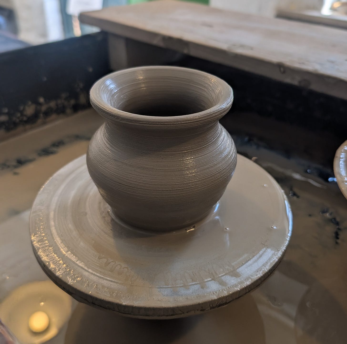 A small, freshly thrown clay pot sits on a pottery wheel in a ceramics studio. The wet grey clay has a rounded body, narrow neck and gently flared rim, with fine throwing lines visible around its surface. The background is softly blurred, drawing attention to the handmade pot as it rests on the wheel, symbolising growth, shaping and becoming. A small, freshly thrown clay pot sits on a pottery wheel in a ceramics studio. The wet grey clay has a rounded body, narrow neck and gently flared rim, with fine throwing lines visible around its surface. The background is softly blurred, drawing attention to the handmade pot as it rests on the wheel, symbolising growth, shaping and becoming.