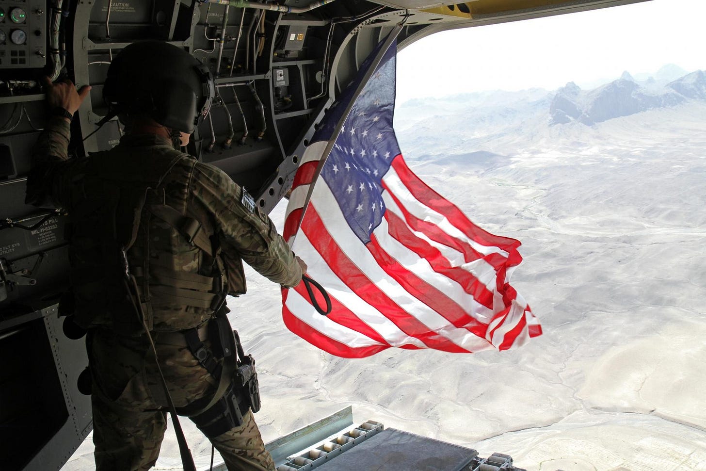 Photo of American soldier holding a flag over an open plane door high above the earth. 