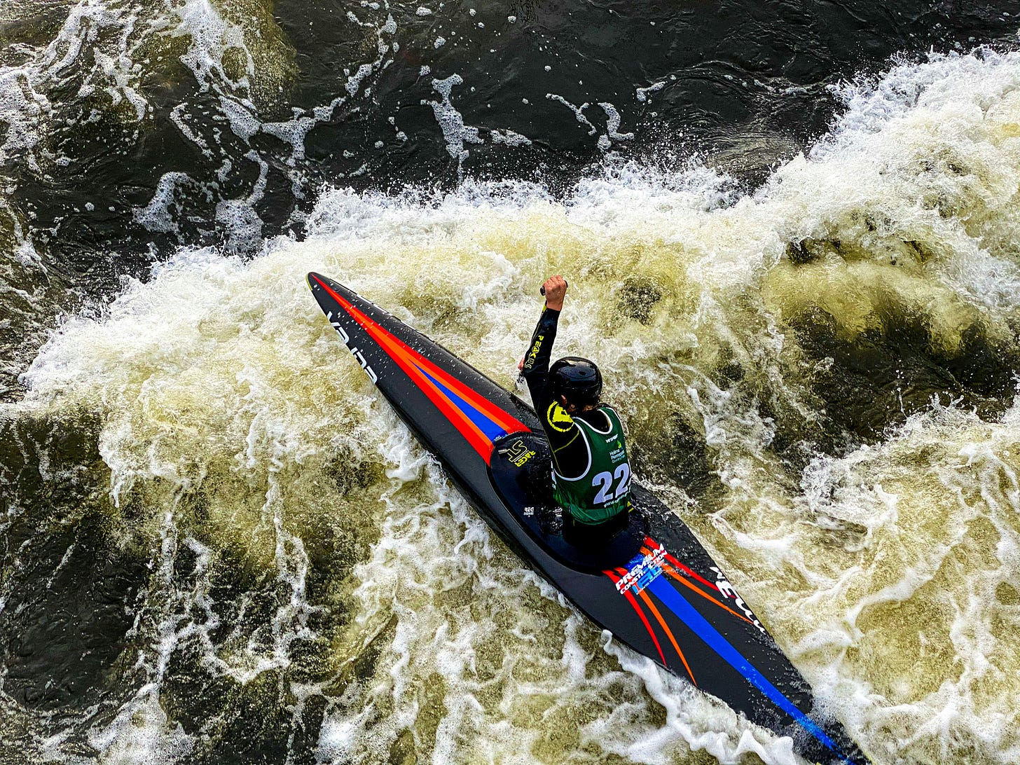 An athlete in a black and green suit is in a black red and blue kayak, paddling through whitewater.