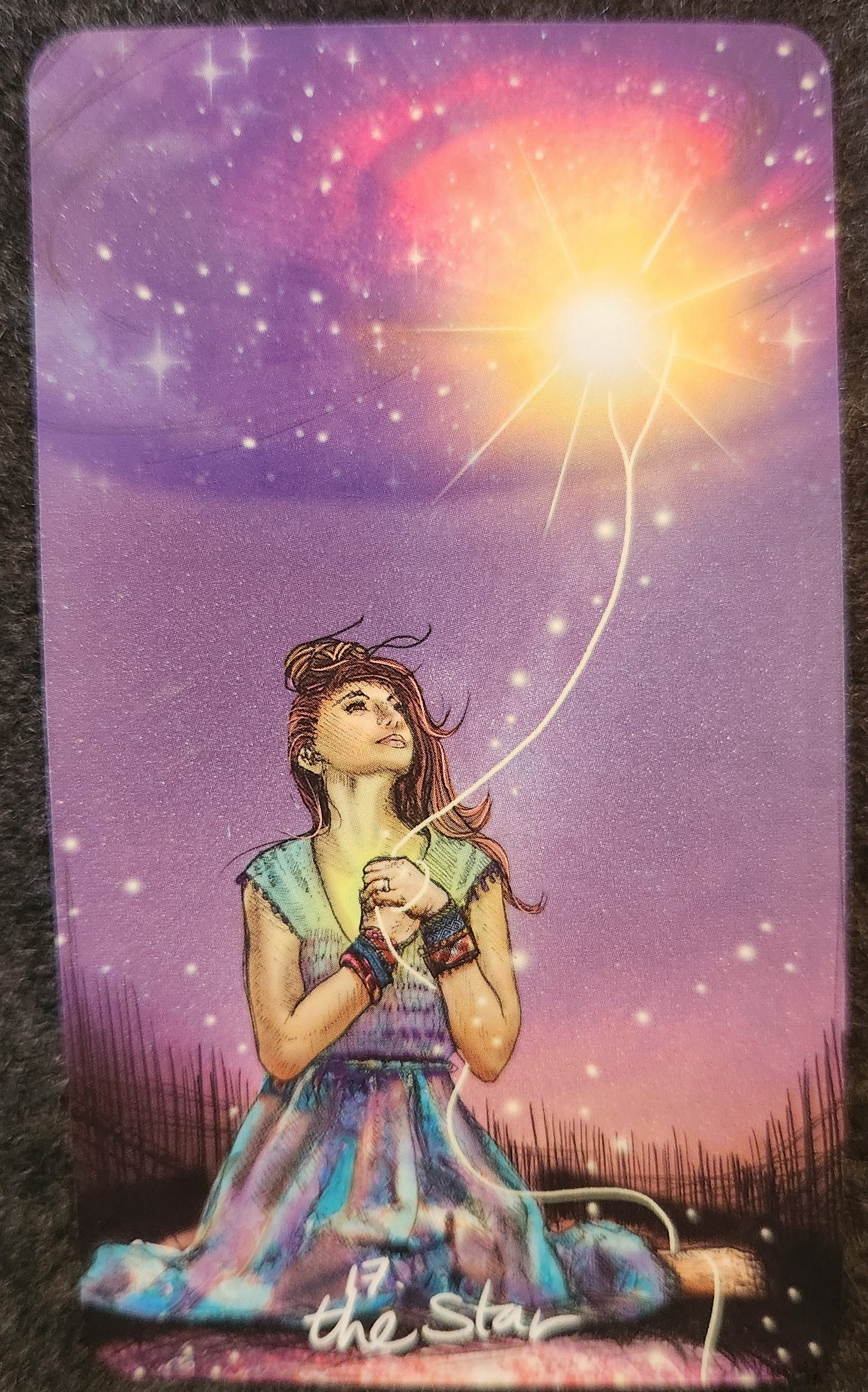 A tarot card labelled 16. The Star. A woman in a multicoloured dress is kneeling in a meadow or field. She is clinging to a string that seems to be connected to a brightly shining star in the sky. She is looking up at the star.