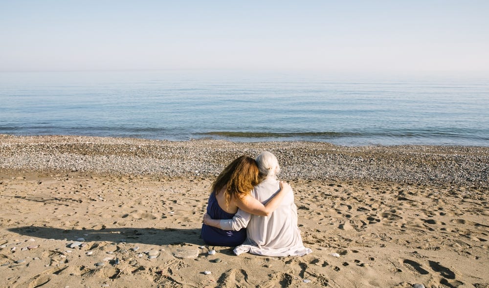 Rear view of elderly mother and adult child holding each other on a beach, looking out at calm waters.