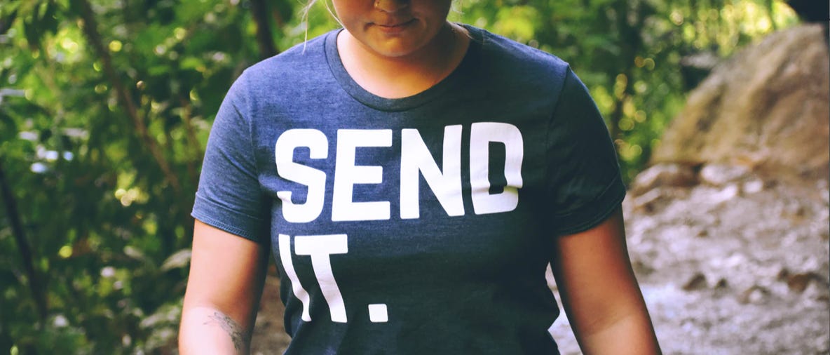 A woman writer wearing a Tshirt saying "send it." walks through a forested area.