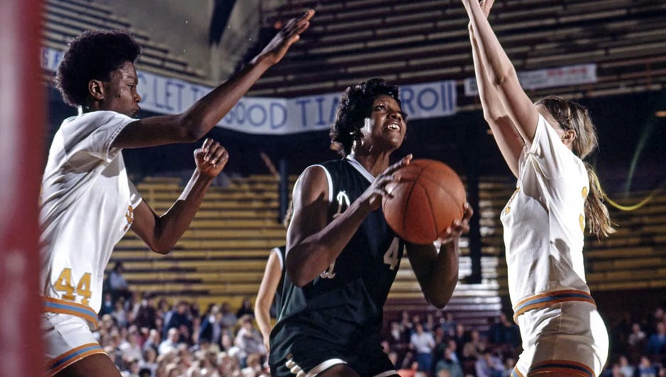Three women’s basketball players in the middle of a play; Lusia Harris, in the middle, is wearing a black uniform and has the ball, looking up as if about to take a shot, while two women in white uniforms on either side of her have their arms up guarding her