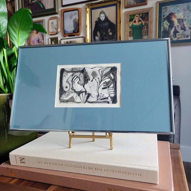 May include: An abstract black and white artwork in a silver frame with a light blue mat. The artwork is displayed on a small gold stand, resting on a stack of books. The background features a collection of framed pictures.