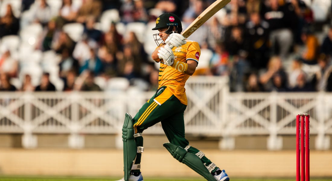A cricket player wearing yellow and green kit swinging his wooden bat in front of a crowd