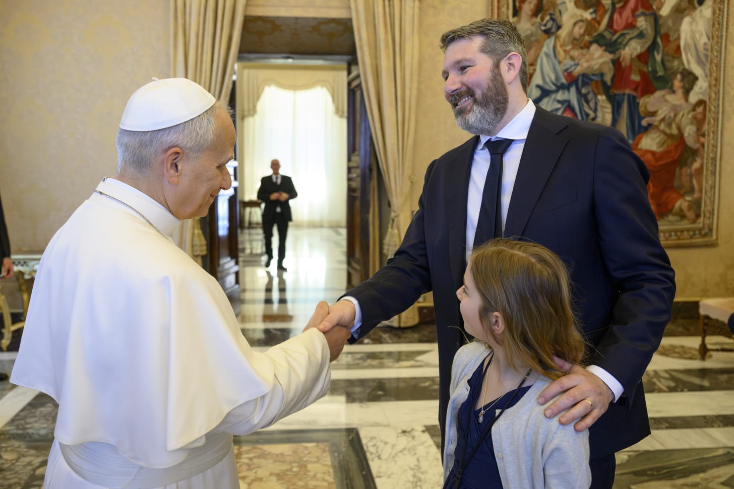 A bearded man in a dark suit and tie shakes hands with a person in white papal vestments and a white zucchetto in an ornate room with tapestries, marble floors, and gold curtains. A young girl stands between them, looking up at the Pope. A security guard stands in a doorway in the background.