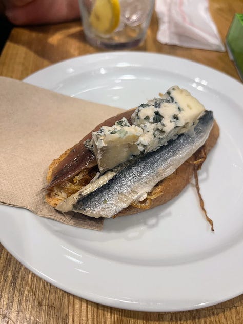 anchovies and blue cheese on a bed of fried onion, a bar meat station with hanging jamon and wrapped cheese, pickled peppers, olives and anchovies on a toothpick