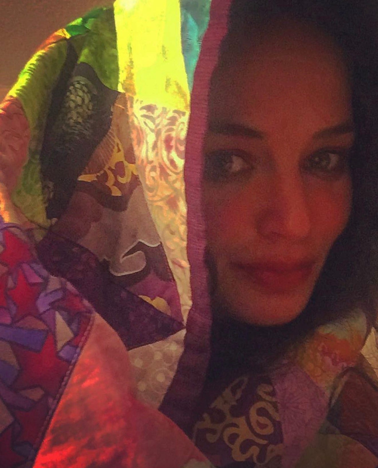 A close-up, warm-toned portrait of Meredith Yayanos looking slightly off-camera. Her head and shoulders are draped in a vibrant, multicolored patchwork quilt featuring various patterns, including stars and paisley, which acts as a hood and casts soft shadows across her face. A close-up, warm-toned portrait of Meredith Yayanos looking slightly off-camera. Her head and shoulders are draped in a vibrant, multicolored patchwork quilt featuring various patterns, including stars and paisley, which acts as a hood and casts soft shadows across her face.