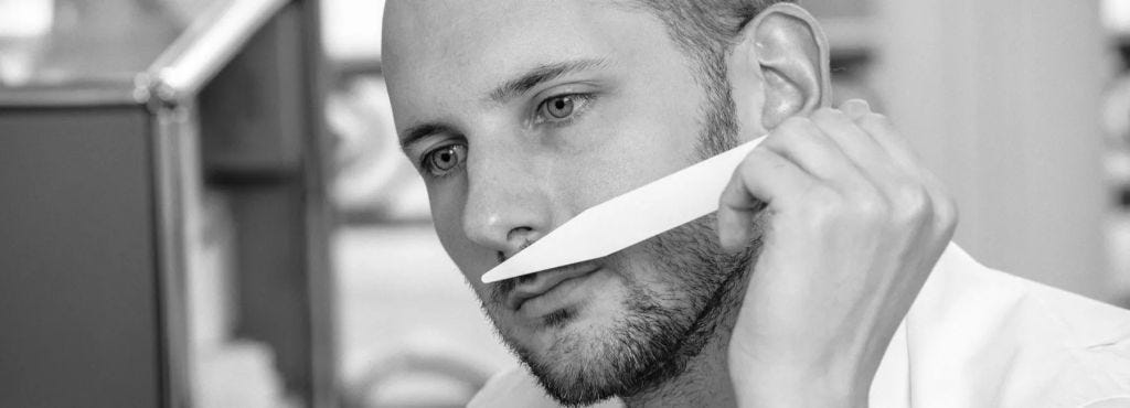 A man with a beard thoughtfully holding a strip of paper near his mouth while looking intently at it.