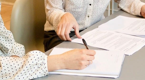 woman signing on white printer paper beside woman about to touch the documents woman signing on white printer paper beside woman about to touch the documents