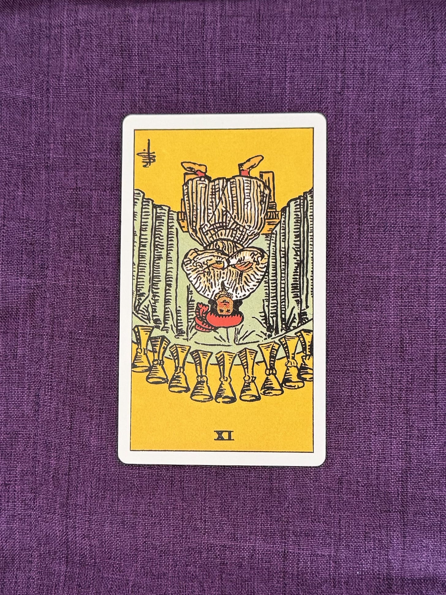 A tarot Card is laid on a purple linen cloth, the card depicts: A middle-aged man is sitting on a wooden bench while his arms are crossed, and his face are showing quite a level of contentment. He is dressed elegantly with a red head-dress. At his background, you will notice there are nine golden cups, structured and arranged in an orderly manner.