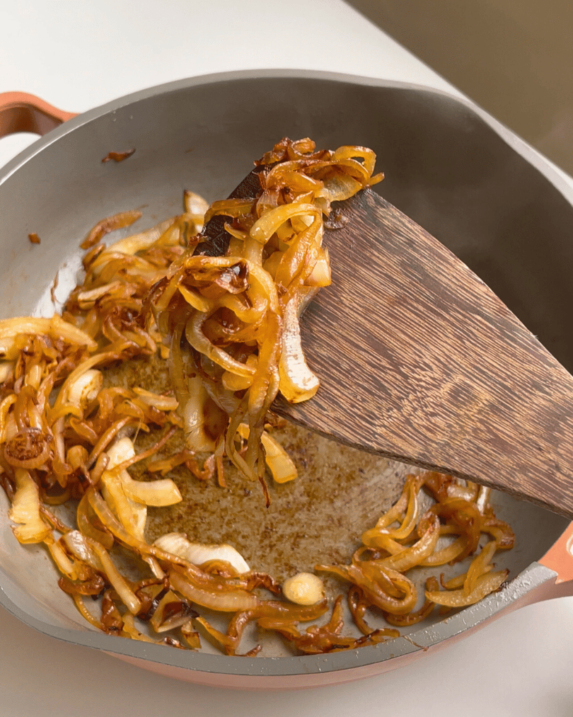 caramelized onions in a grey pan with a wooden spoon holding some onions up higher