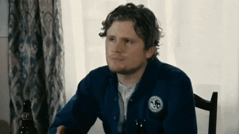 Man in blue jacket sits at a table with bottles, looking contemplative. White curtains and patterned drapes in the background.