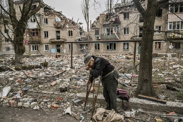 A man leaning on crutches in a street filled with debris, with a destroyed building behind him.