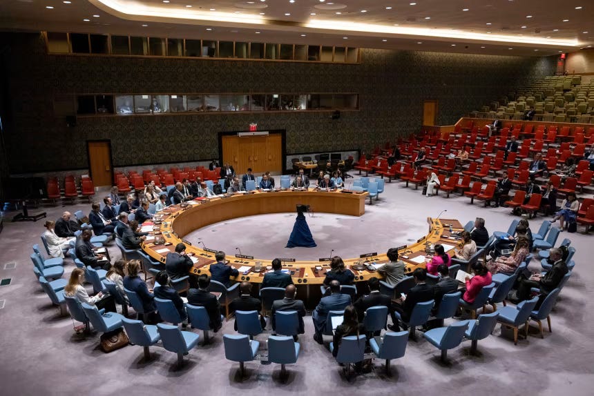The Security Council chamber seen from above during a session, delegates seated around the horseshoe table with rows of blue and red seats behind and a mural on the far wall. The Security Council chamber seen from above during a session, delegates seated around the horseshoe table with rows of blue and red seats behind and a mural on the far wall.