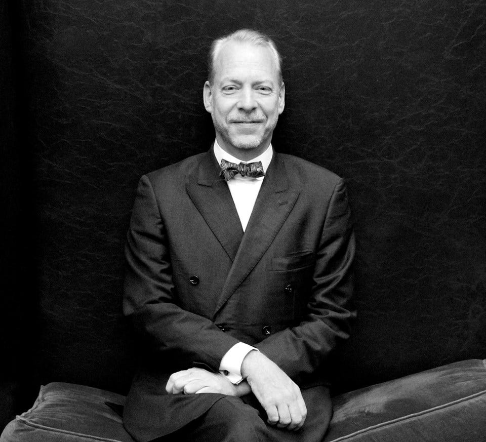 Black and white portrait of Jeffrey Tucker seated in a double breasted suit and bow tie, smiling. Black and white portrait of Jeffrey Tucker seated in a double breasted suit and bow tie, smiling.
