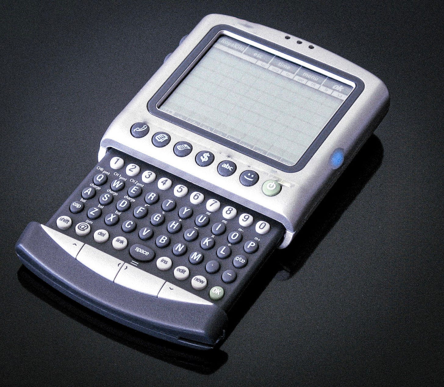 An old-style handheld PDA, with a small blank screen at the top and numerous buttons on a pull-out alphanumeric keyboard beneath the screen. The keyboard also includes various function keys labeled "shift," "@," "link," etc. Although the image is mostly drained of color, on the right edge of the PDA is a bright blue button. An old-style handheld PDA, with a small blank screen at the top and numerous buttons on a pull-out alphanumeric keyboard beneath the screen. The keyboard also includes various function keys labeled "shift," "@," "link," etc. Although the image is mostly drained of color, on the right edge of the PDA is a bright blue button.