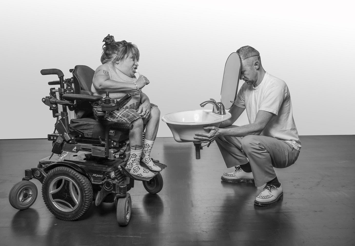 A black and white photo of a femme in a powerchair brushing her teeth at a sink and mirror held up by a white man.