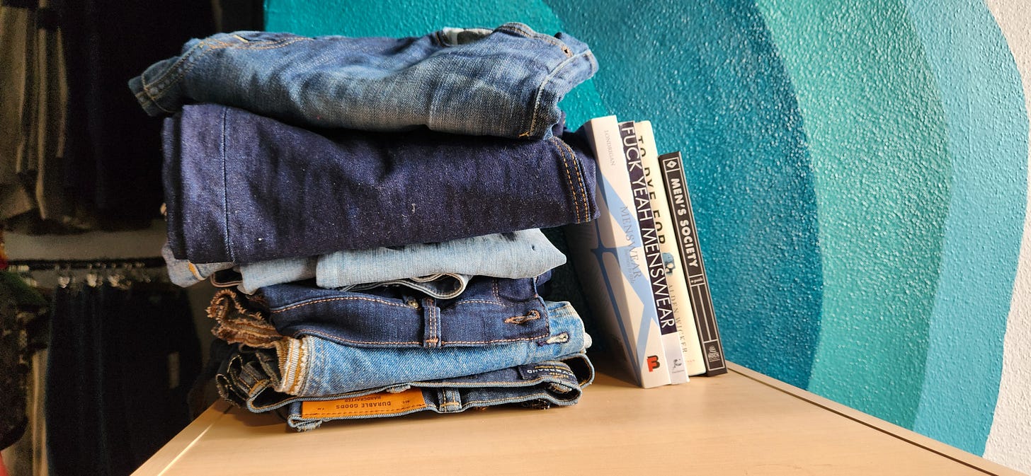 A stack of various blue denim clothing sits on top of a shelf next to 4 books in front of a teal monochromatic wave mural.