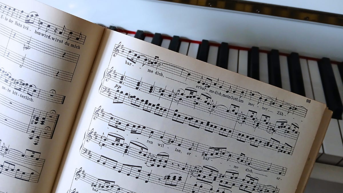 An open sheet music book with German lyrics rests on the keys of a piano, showing musical notes and staff lines. The piano keyboard is partially visible in the background. An open sheet music book with German lyrics rests on the keys of a piano, showing musical notes and staff lines. The piano keyboard is partially visible in the background.