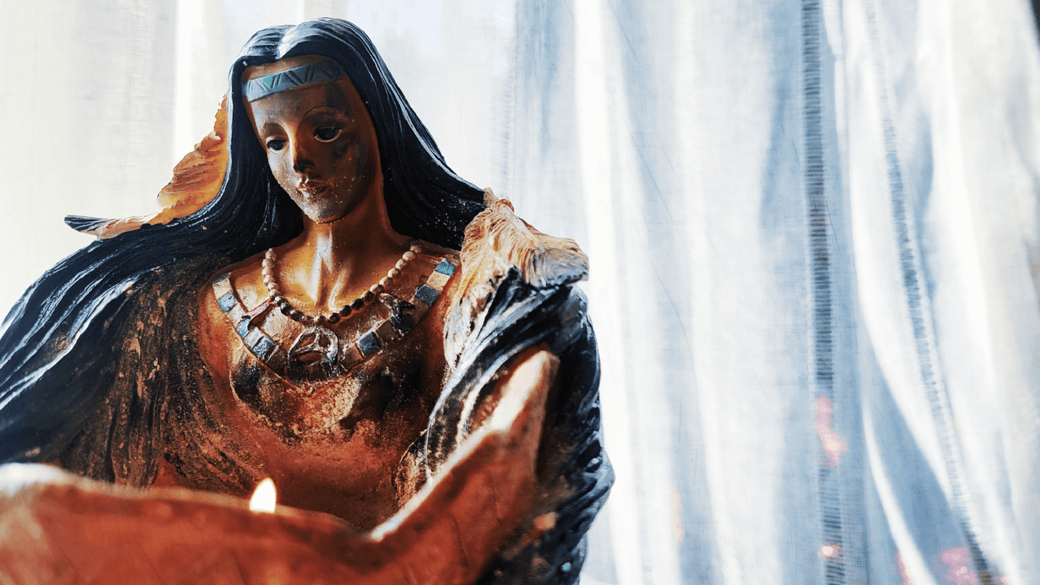 candle held my a nurturing indigenous woman. back drop is a sheer white curtain in the filtered light. Her face is ash covered, solemn but approachable.