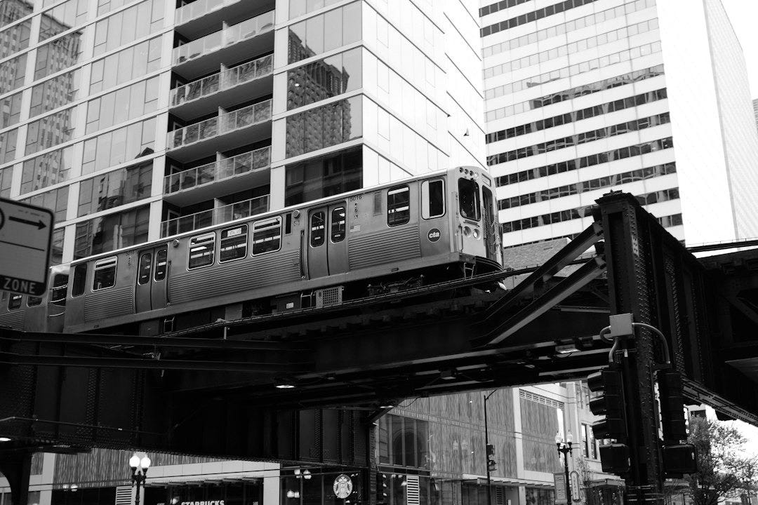 a silver train traveling over a bridge next to tall buildings a silver train traveling over a bridge next to tall buildings