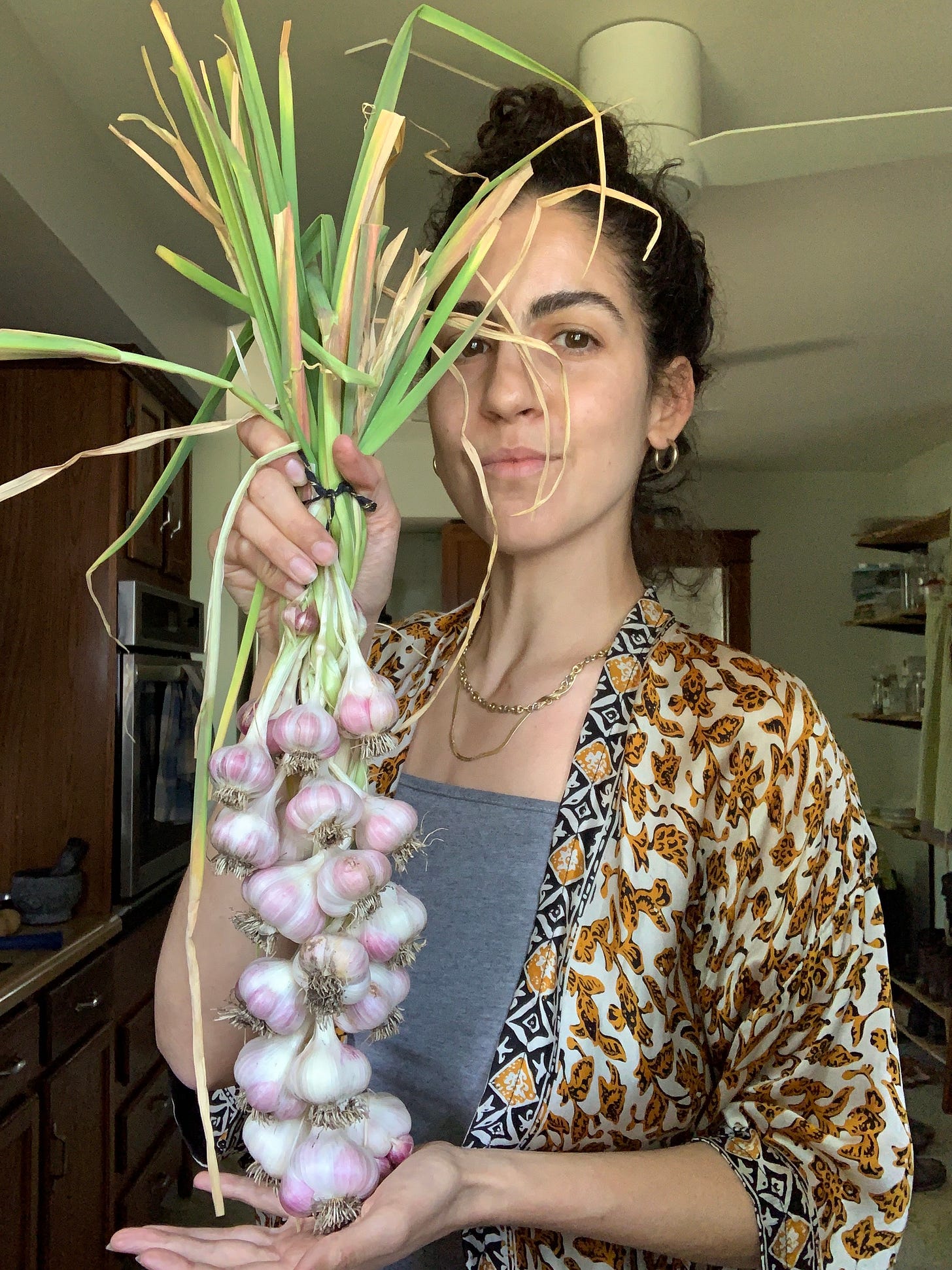 A person with olive skin and dark hair pulled into a bun is holding freshly dug garlic bulbs that have been made into a braid looking proud. She's standing in a dark room and wearing a silk kimono style top over a gray tank top.