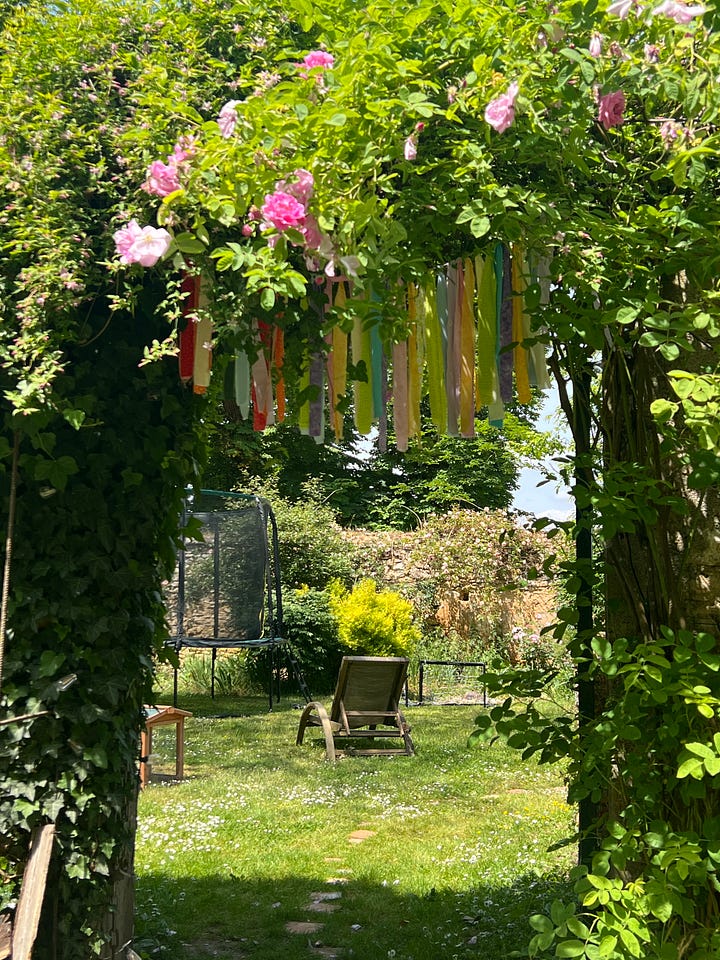 Four photos of a walled French garden in spring: a canopy tent on the grass under blossoming trees, a wooden playhouse and swing set beneath a cherry tree in full pink bloom, a young girl planting seedlings in a raised bed beside a wheelbarrow, and a trampoline framed by climbing roses and a ribbon archway.
