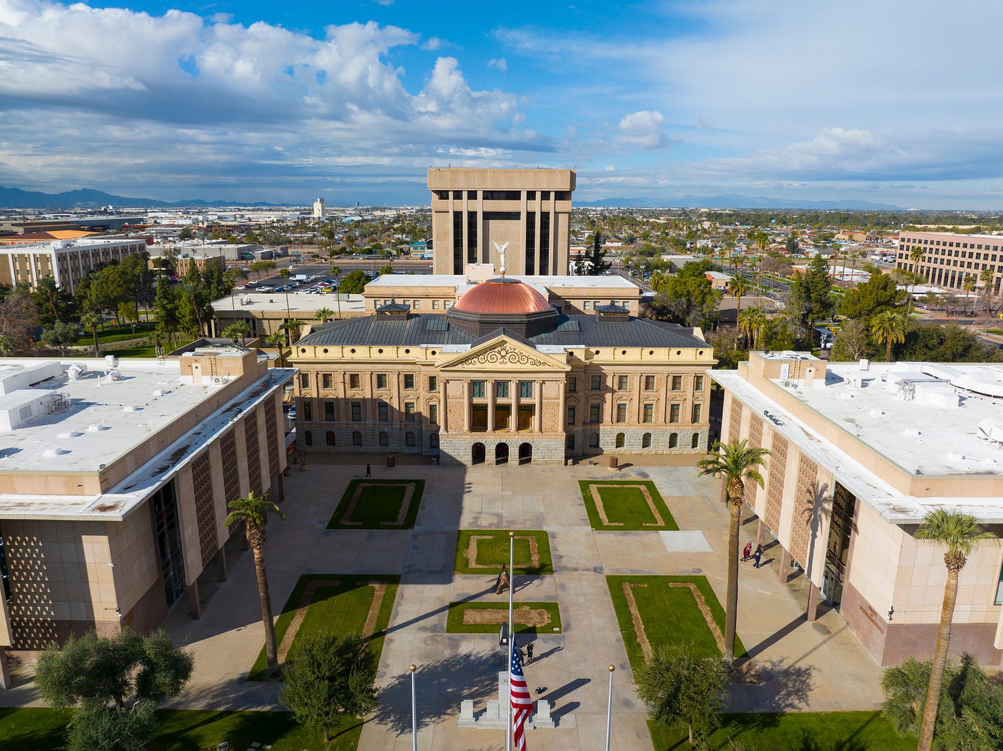 Aerial view of the Arizona State Capitol complex in Phoenix, with the historic domed capitol building centered between two modern legislative buildings and a courtyard lined with palm trees. Aerial view of the Arizona State Capitol complex in Phoenix, with the historic domed capitol building centered between two modern legislative buildings and a courtyard lined with palm trees.