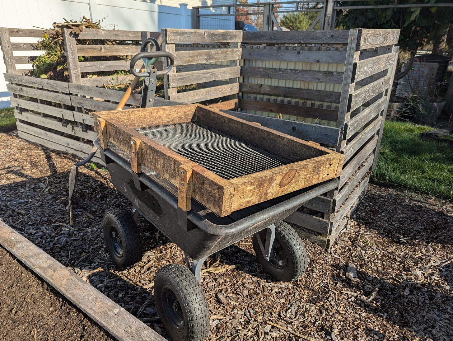 A garden cart with a sifter on top is parked in front of a compost structure in the garden on a sunny day.  The sifter is a wooden frame with 1/2" mesh hardware cloth attached to the bottom.