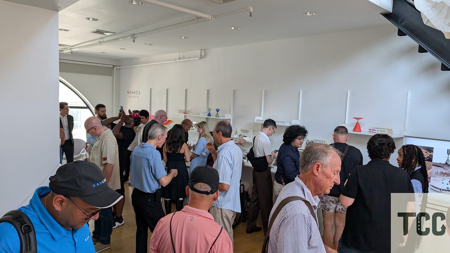 Dozens of attendees at a NOMOS event closely inspect watches displayed on minimalist white shelves. Some are photographing watches, while others are chatting or browsing.