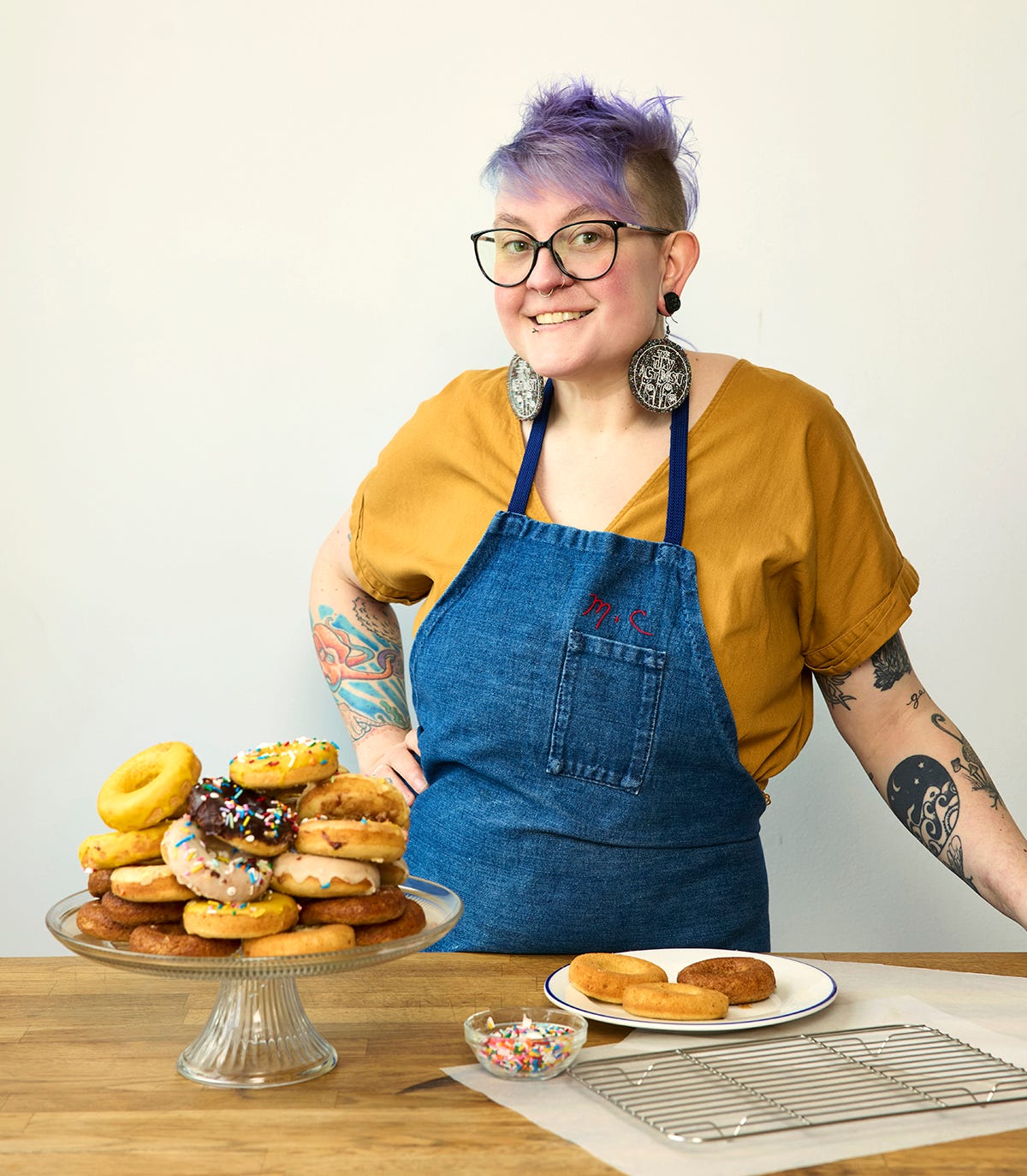 Corrie is a white human with short purple hair, tattoos, and glasses. They are smiling at the camera, wearing a denim apron over a yellow shirt. They are standing at a table or counter. There are frosted donuts of different varieties on a cake stand, and some baking supplies on the counter. Corrie is a white human with short purple hair, tattoos, and glasses. They are smiling at the camera, wearing a denim apron over a yellow shirt. They are standing at a table or counter. There are frosted donuts of different varieties on a cake stand, and some baking supplies on the counter.